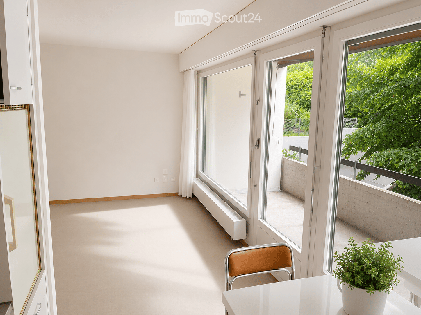 Bright, minimalist room with large windows and sliding glass doors leading to a balcony. Features a white table, a single chair, and a potted plant on the windowsill.