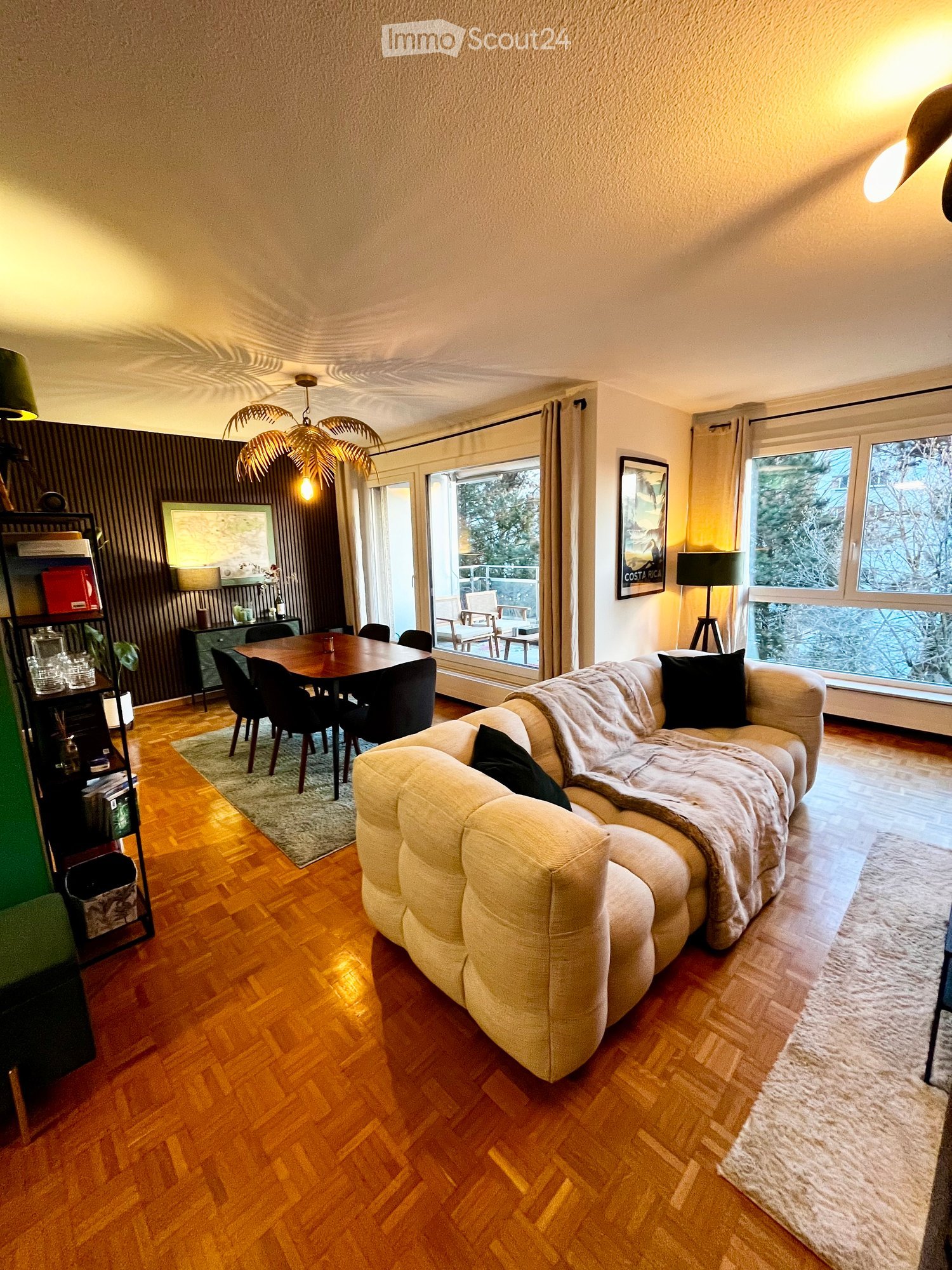 Furnished Room. A modern living room with a large sectional sofa, a dining table with chairs, a floor lamp, a shelf with decorative items, and large windows with a view of the outdoors.