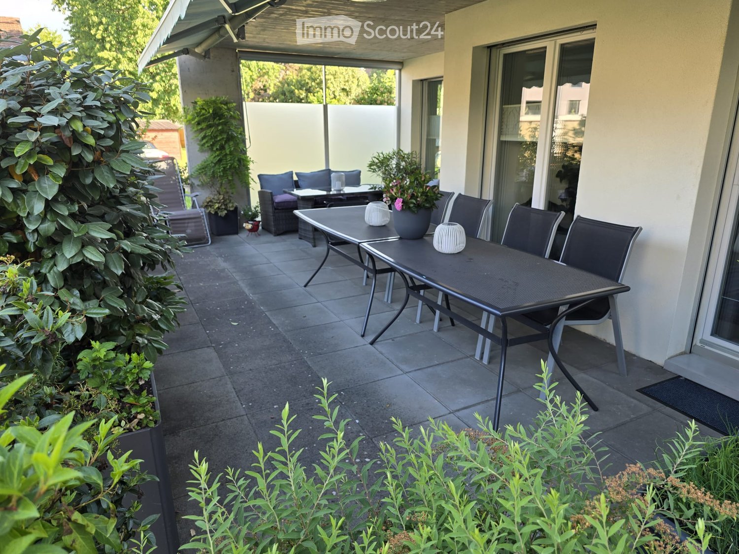Balcony with a dining table, chairs, and potted plants, surrounded by greenery and a modern building.