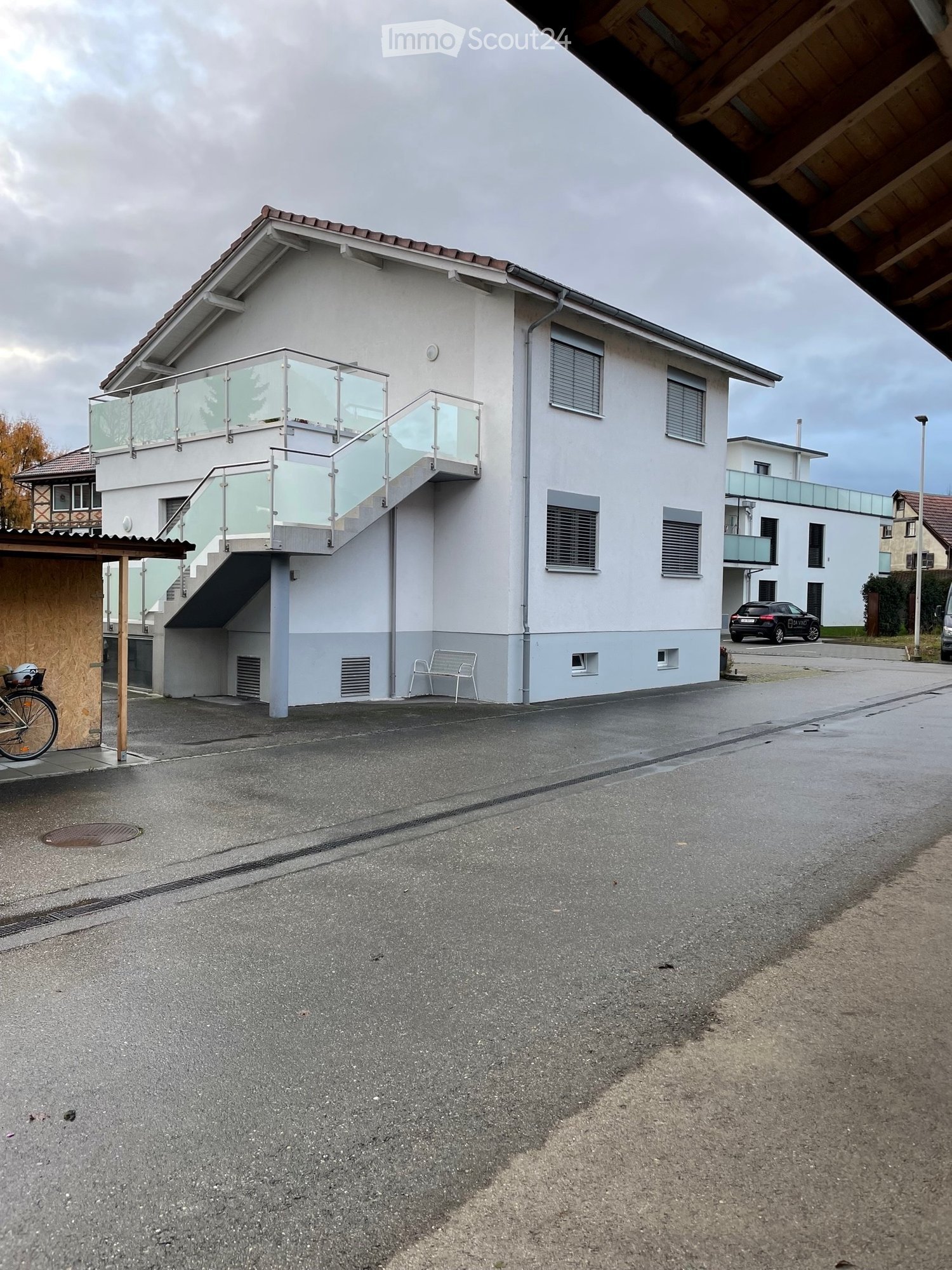 Two-story house with white exterior, glass balcony, and covered entrance area. A bicycle is parked near the entrance, and a bench is visible near the base of the house.