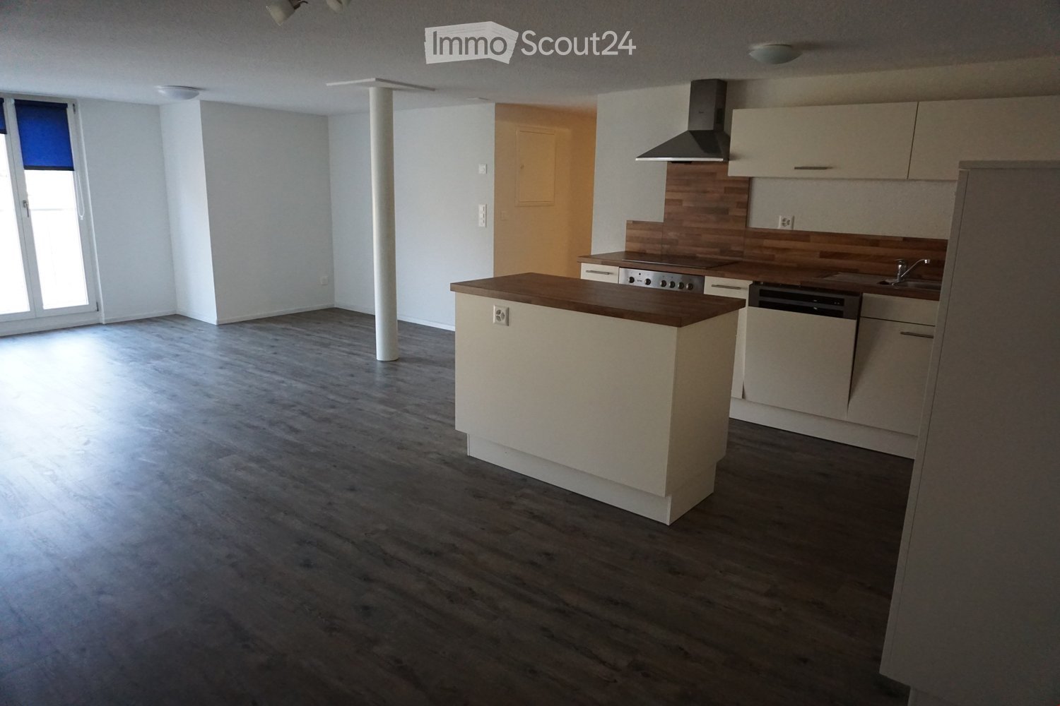 Modern kitchen with light-colored cabinets, a central island, and built-in appliances including a stove, dishwasher, and sink. The space features wooden flooring and ample natural light from a large window.