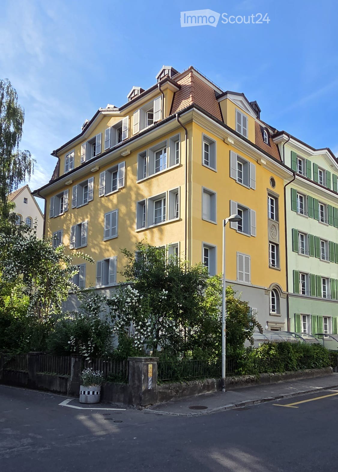 A multi-story yellow building with white shutters and a red-tiled roof, surrounded by greenery and neighboring houses.