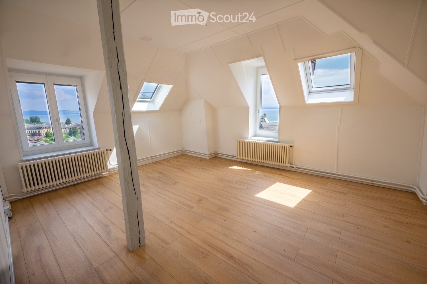 Empty attic room with wooden flooring, white walls, multiple windows, and radiators. Natural light streams in from several windows, including skylights, offering a view of the outdoors.