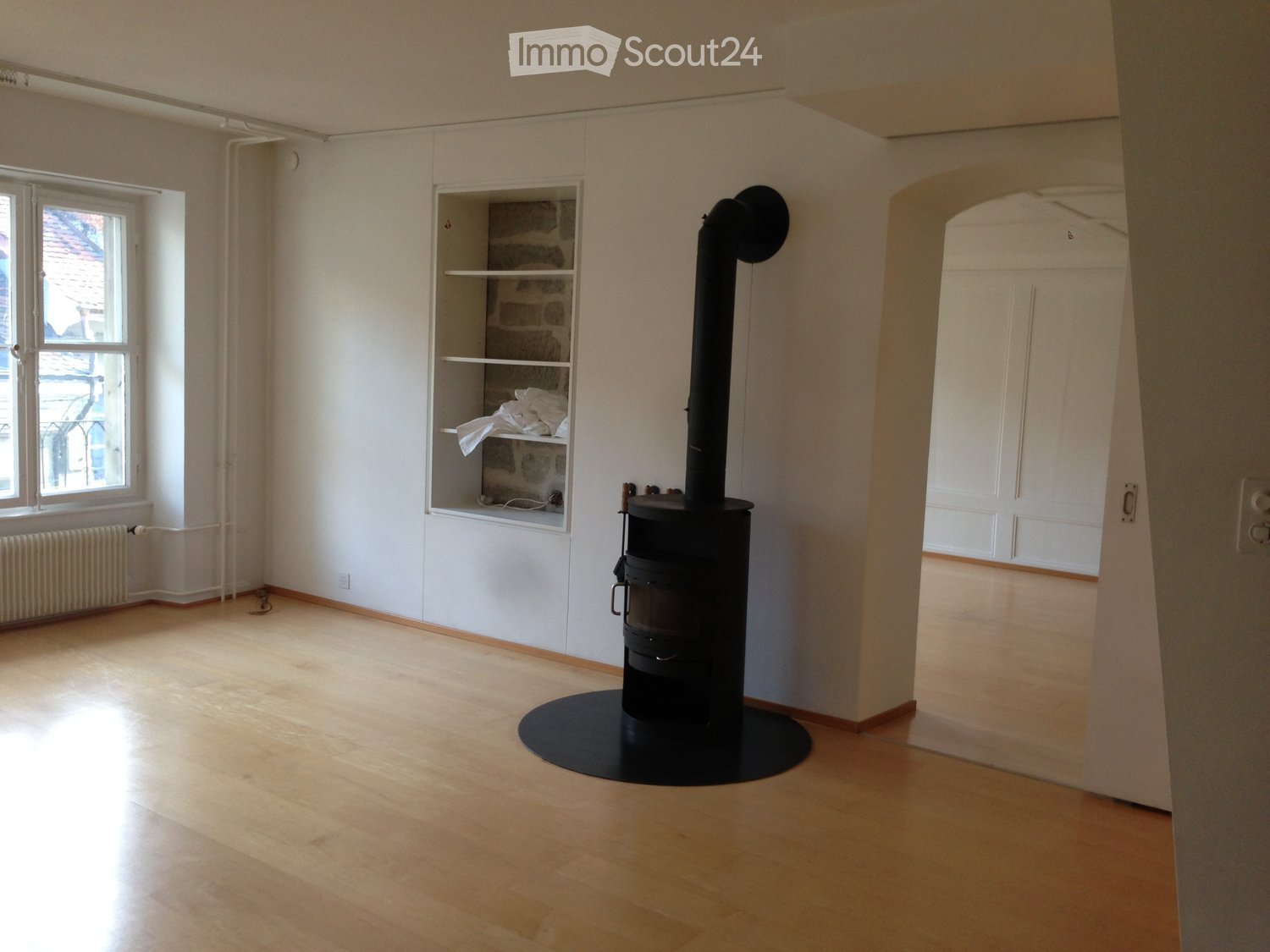 Empty room with wooden flooring, white walls, a black wood-burning stove, a built-in shelf, and a window with a radiator below it.