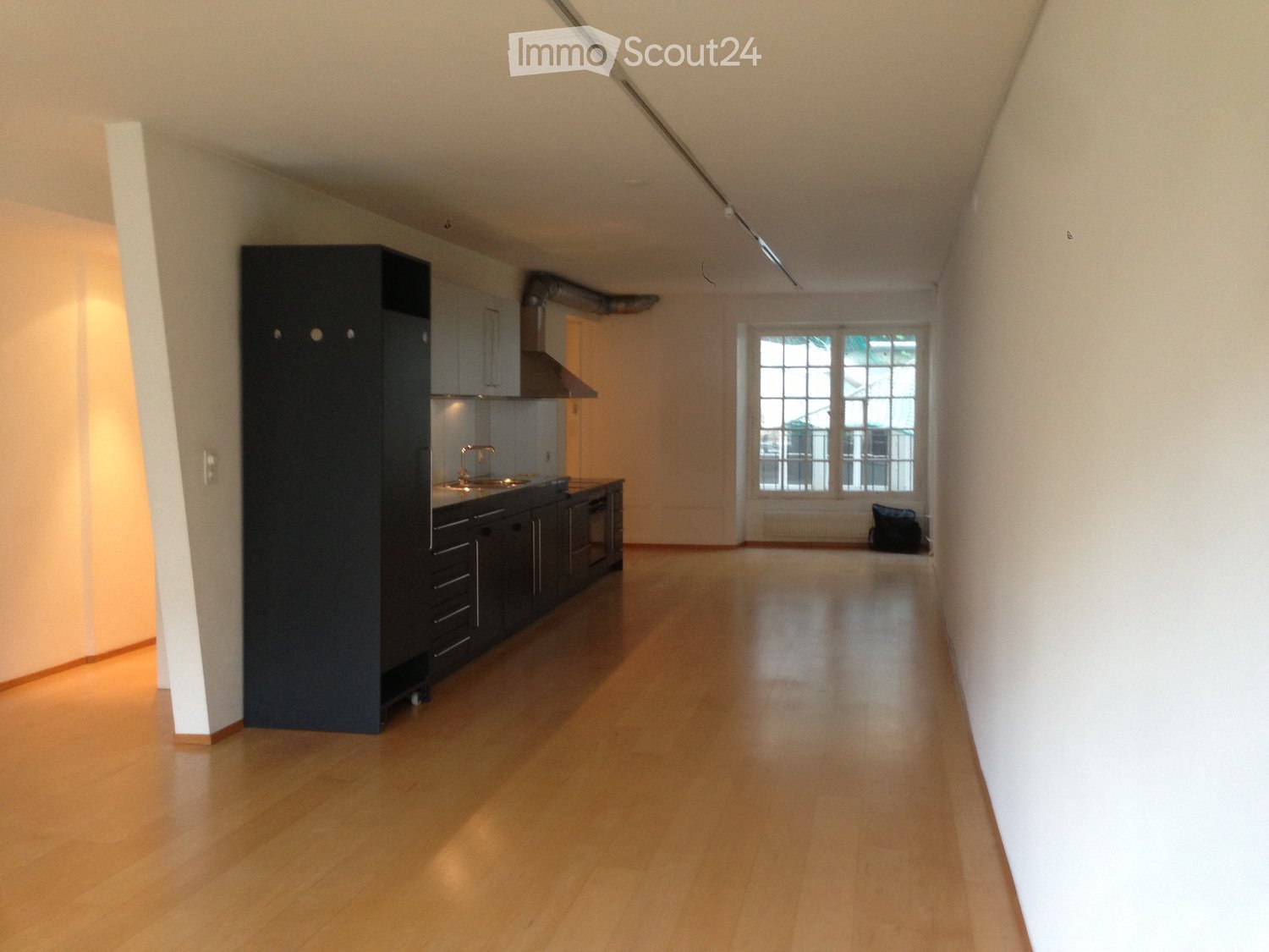 Empty room with wooden flooring, white walls, and a small kitchen area with cabinets and a sink visible in the background.