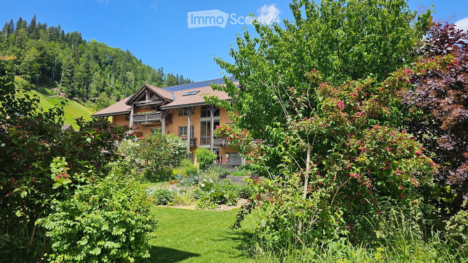 A two-story house with a tiled roof, surrounded by lush greenery and a well-maintained garden. The house features large windows and a balcony on the upper floor.