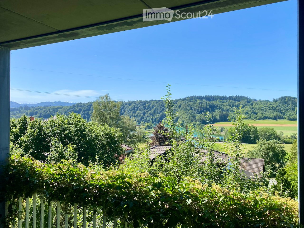 Balcony with a view of a lush green landscape, trees, and distant hills under a clear blue sky.