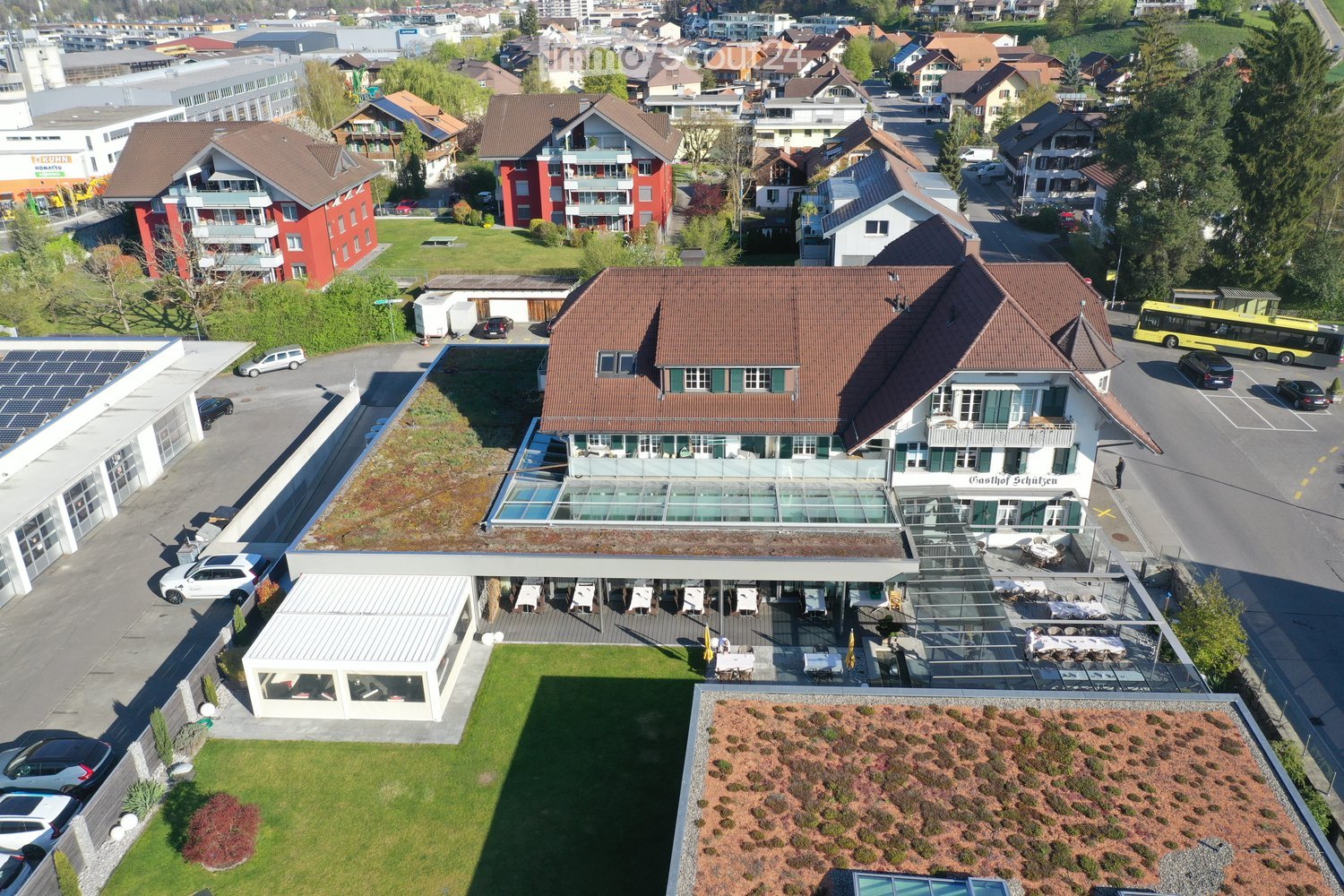 Aerial view of a multi-story building with a red-tiled roof, surrounded by a well-maintained lawn, outdoor seating area, and parking lot. The building features large windows and a modern design, with additional structures and greenery in the vicinity.