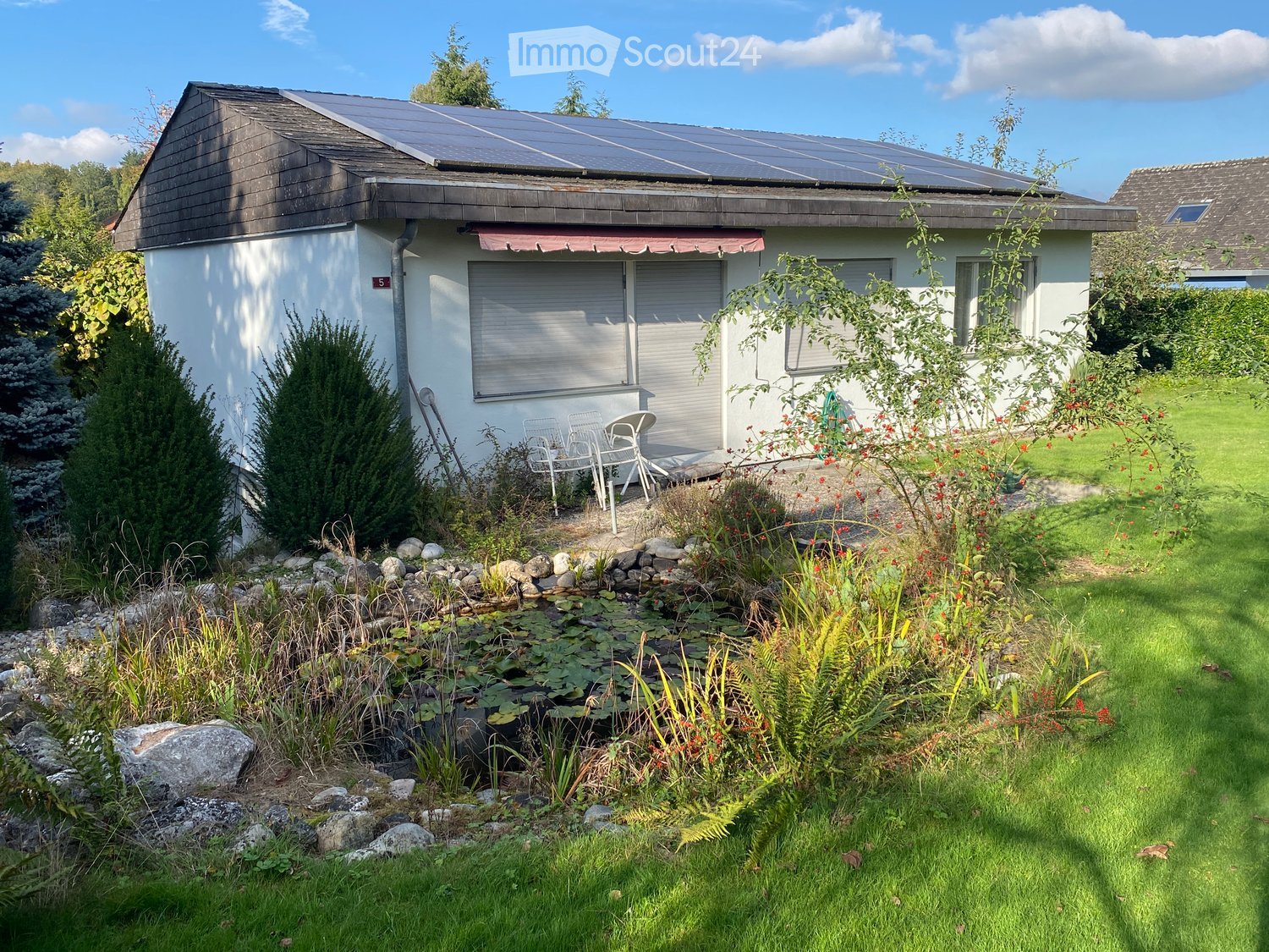 A small white house with solar panels on the roof, surrounded by a well-maintained garden with a pond, rocks, and various plants. There is a patio area with chairs and a table.