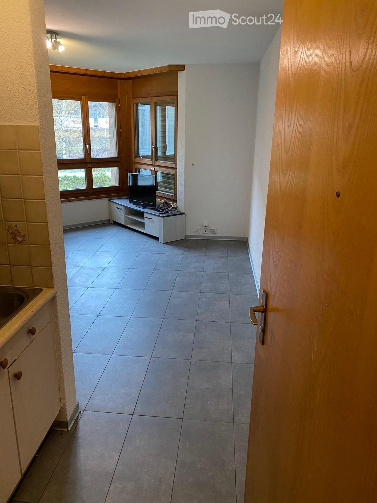 A small, unfurnished room with tiled flooring, a TV stand with a screen, and a sink visible in the background.