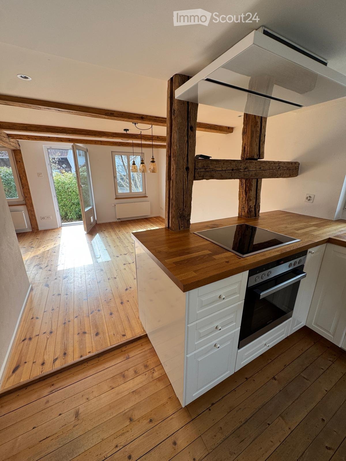 Modern kitchen with wooden countertops, white cabinets, induction stove, and pendant lights. Features exposed wooden beams and a large window with a view of greenery.