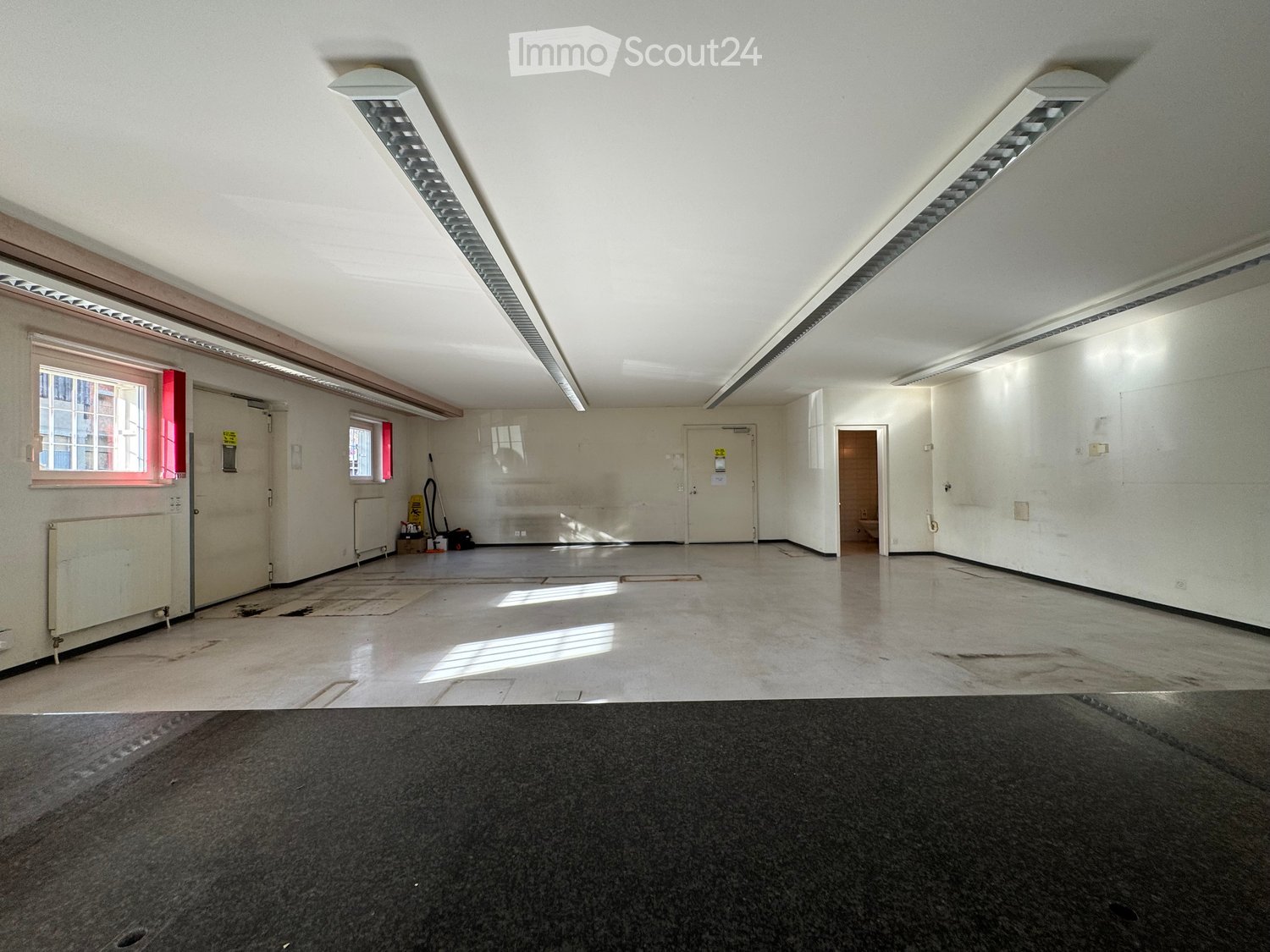 Empty room with white walls, concrete floor, fluorescent lighting, and a few windows. No furniture or equipment present.