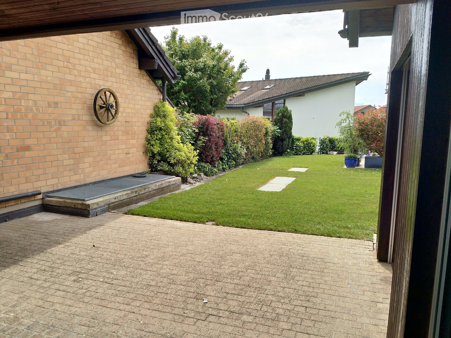 Well-maintained garden with a neatly trimmed lawn, surrounding shrubs, and a paved patio area. A decorative wheel is mounted on the wall of the adjacent building.