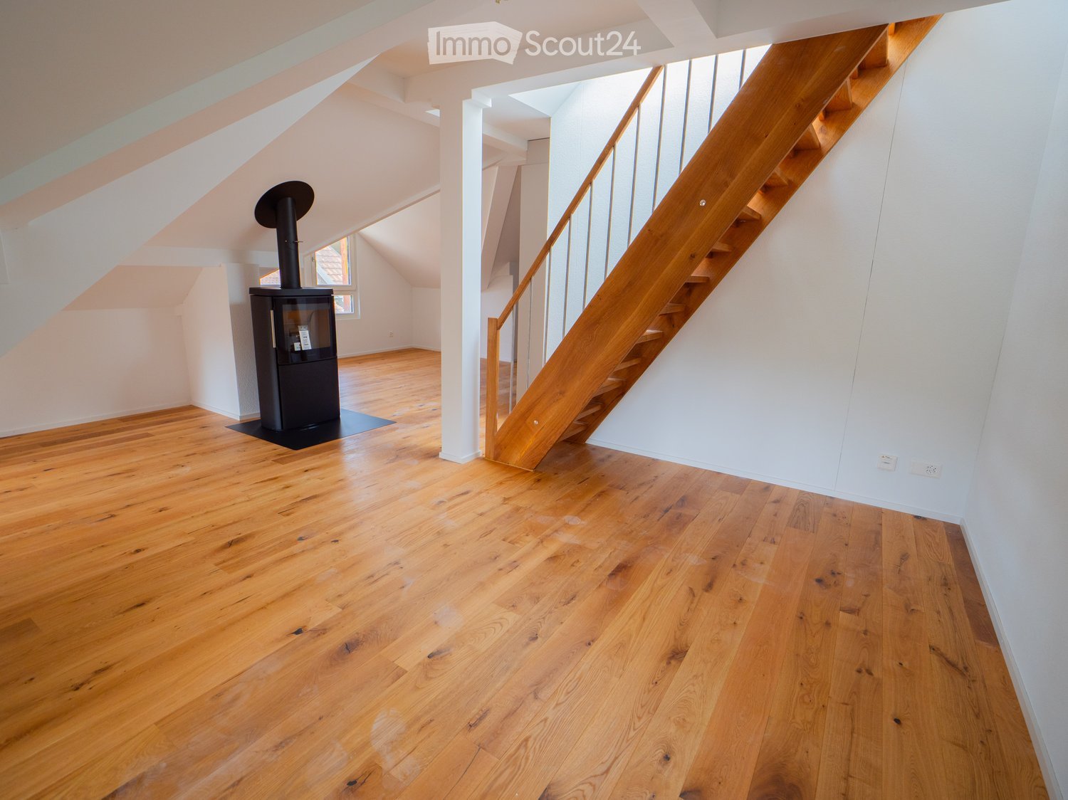 Empty attic space with wooden flooring, white walls, and a staircase leading upwards. A black wood-burning stove is positioned against the wall.
