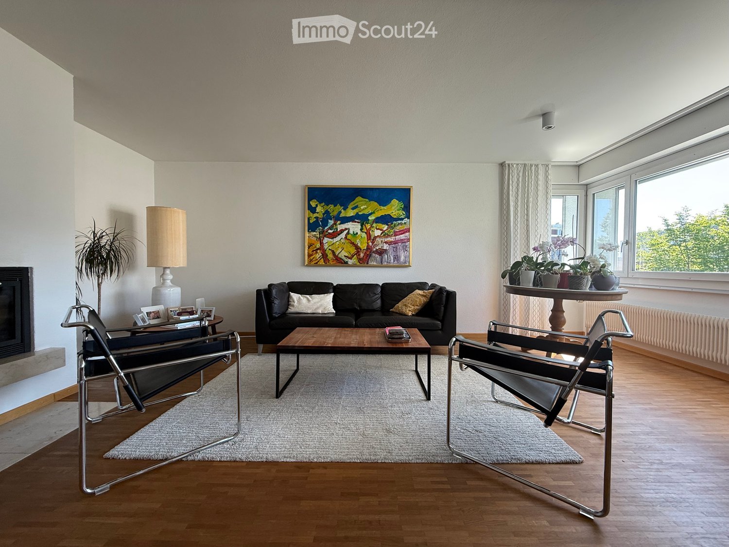 Modern living room with wooden flooring, a black leather sofa, coffee table, two chairs, a side table with a lamp, a fireplace, and a painting on the wall.