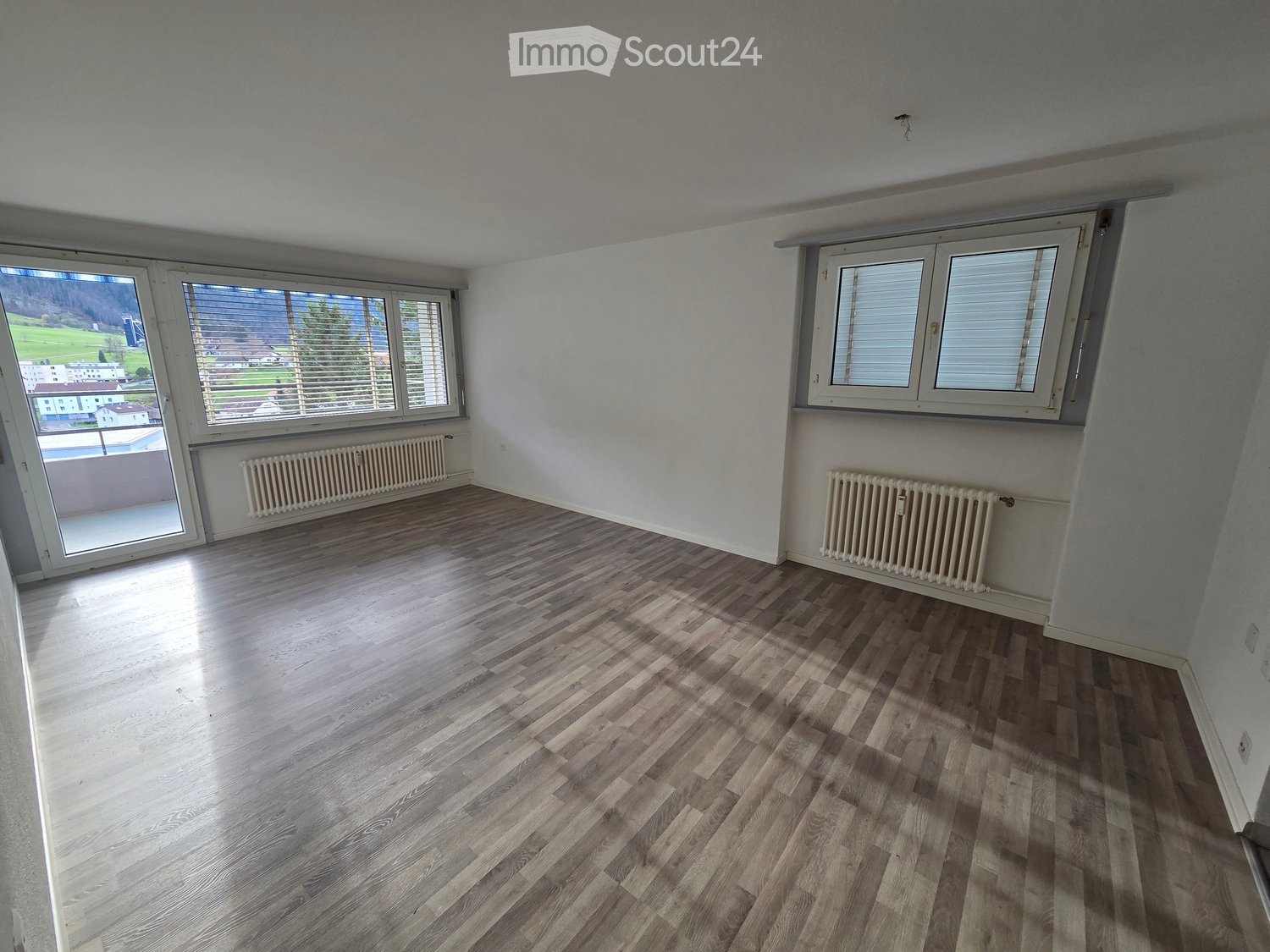 Empty room with wooden flooring, white walls, and large windows with blinds. Radiators are visible along the walls.