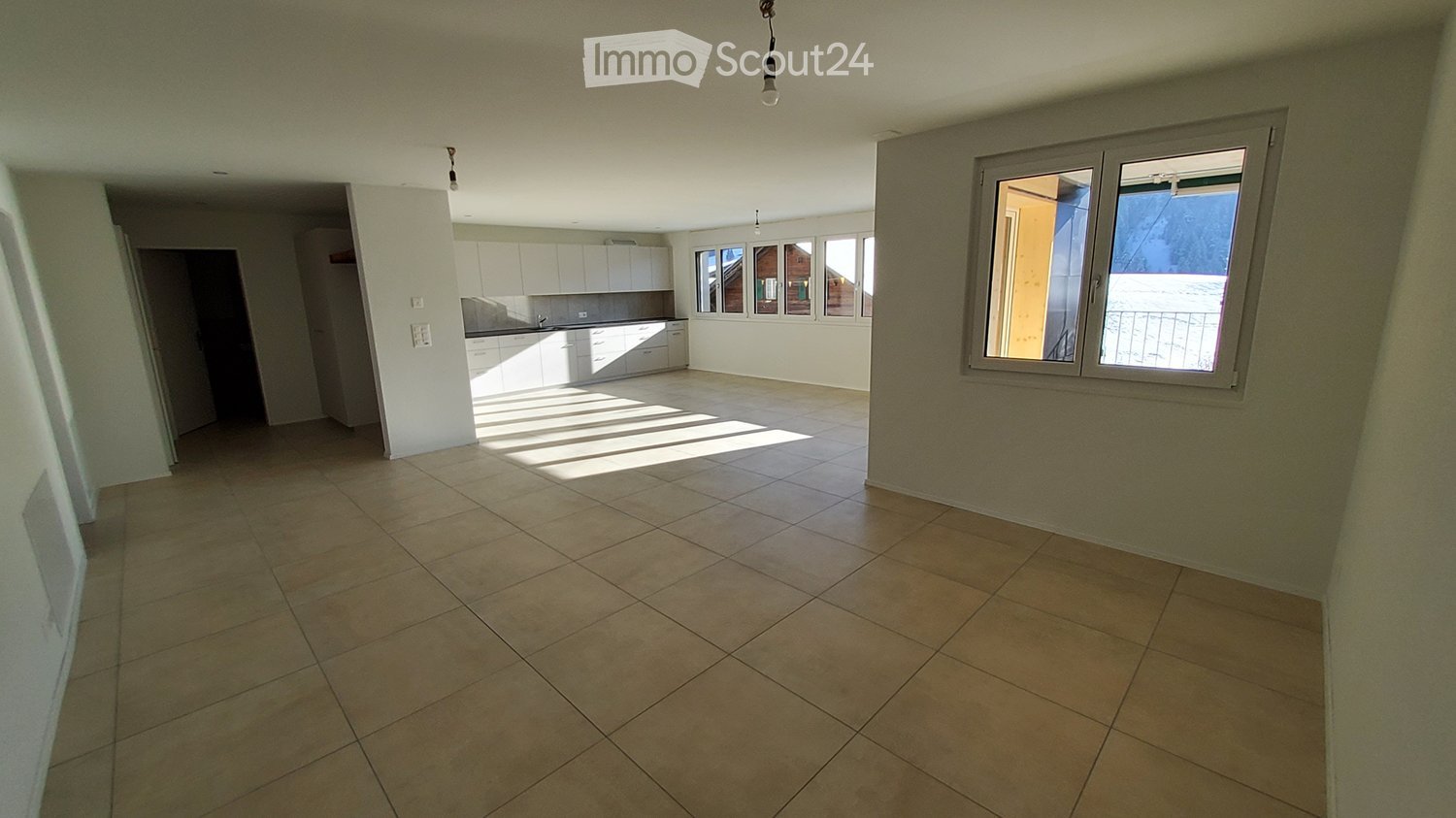 Empty room with tiled floor, white walls, and large windows. The room is connected to a kitchen area with white cabinets and countertops.
