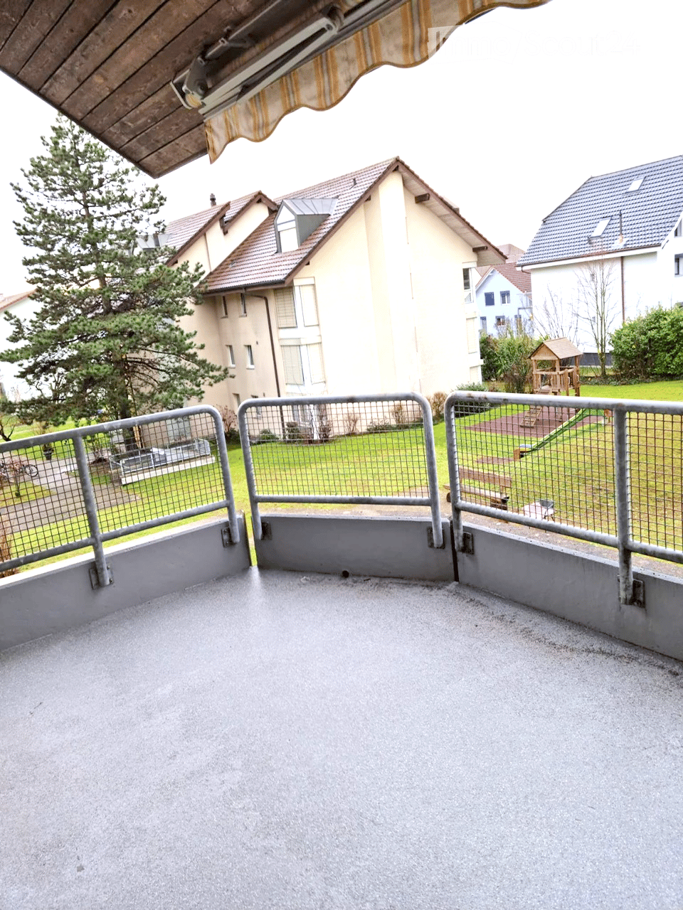Empty balcony with metal railing, concrete floor, and a view of a residential area with houses, trees, and a playground.