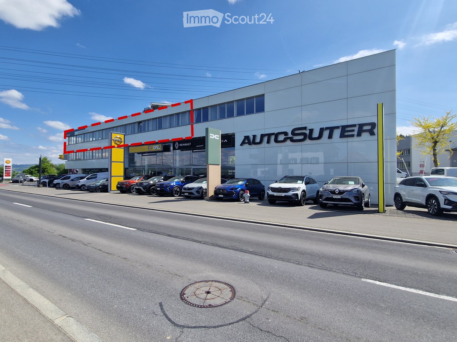 Modern car dealership building with large glass windows and signage for 'AUTOSUTER', several cars parked in front.