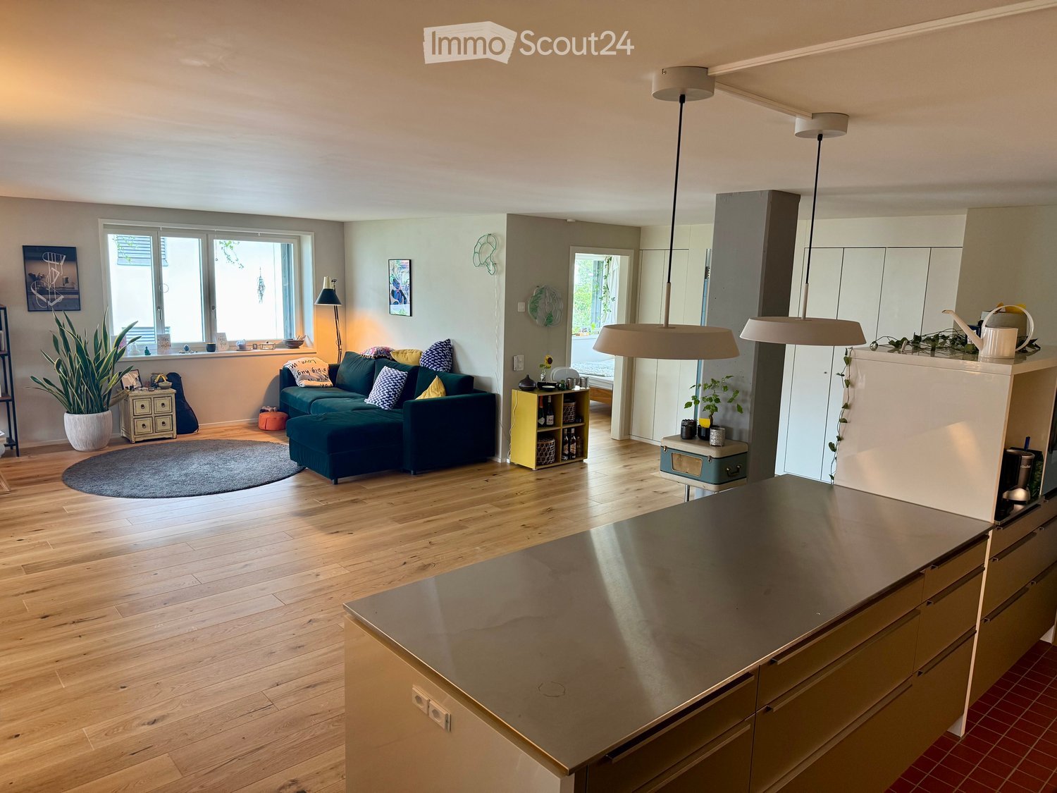Open-plan living and dining area with a green sofa, pendant lights, a kitchen island, and a potted plant. The space features wooden flooring and large windows.