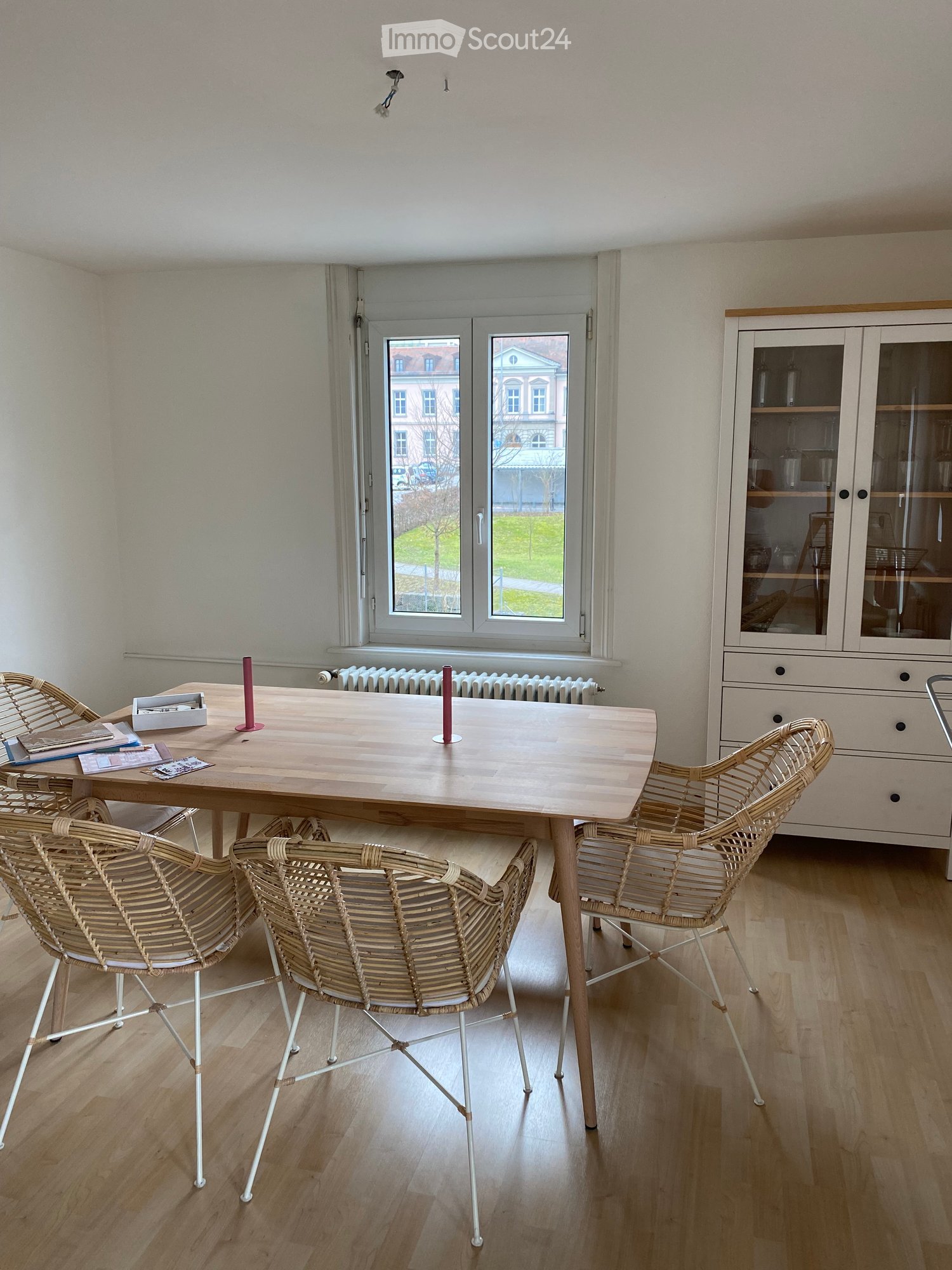 Dining area with a wooden table, four wicker chairs, a white cabinet, and a radiator under a large window.