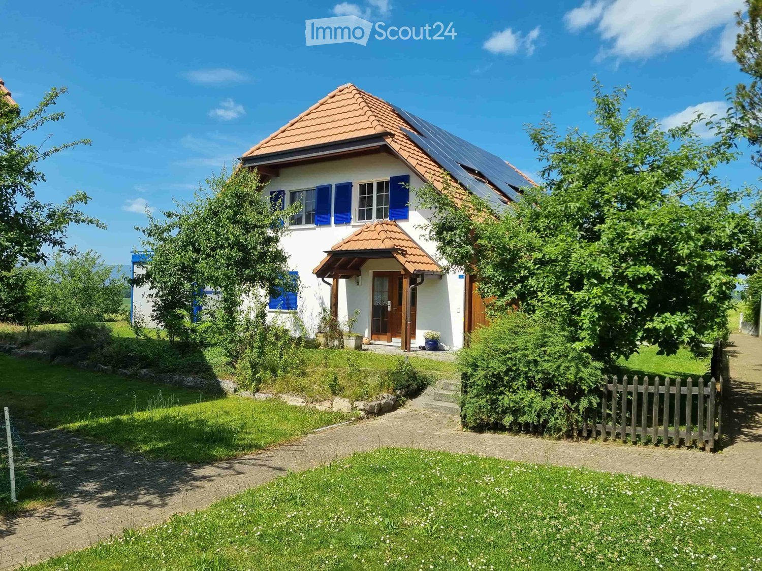 Two-story house with white walls, blue shutters, red-tiled roof, and solar panels on the roof. The house has a small porch with a wooden door and is surrounded by a well-maintained garden with a gravel pathway and wooden fence.