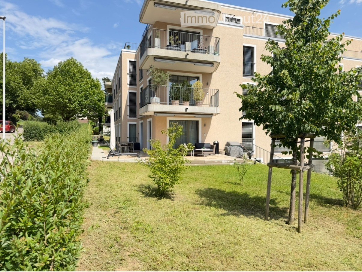 Modern multi-story residential building with balconies, surrounded by a well-maintained garden and outdoor seating area.