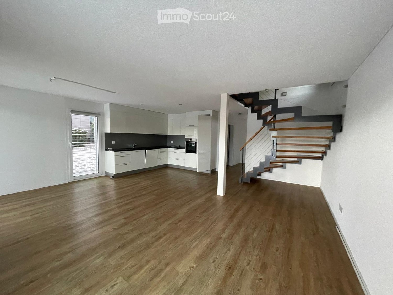 Open-plan space with a modern kitchen area featuring white cabinets and a dark countertop, connected to a living area with wooden flooring and a staircase leading to an upper level.