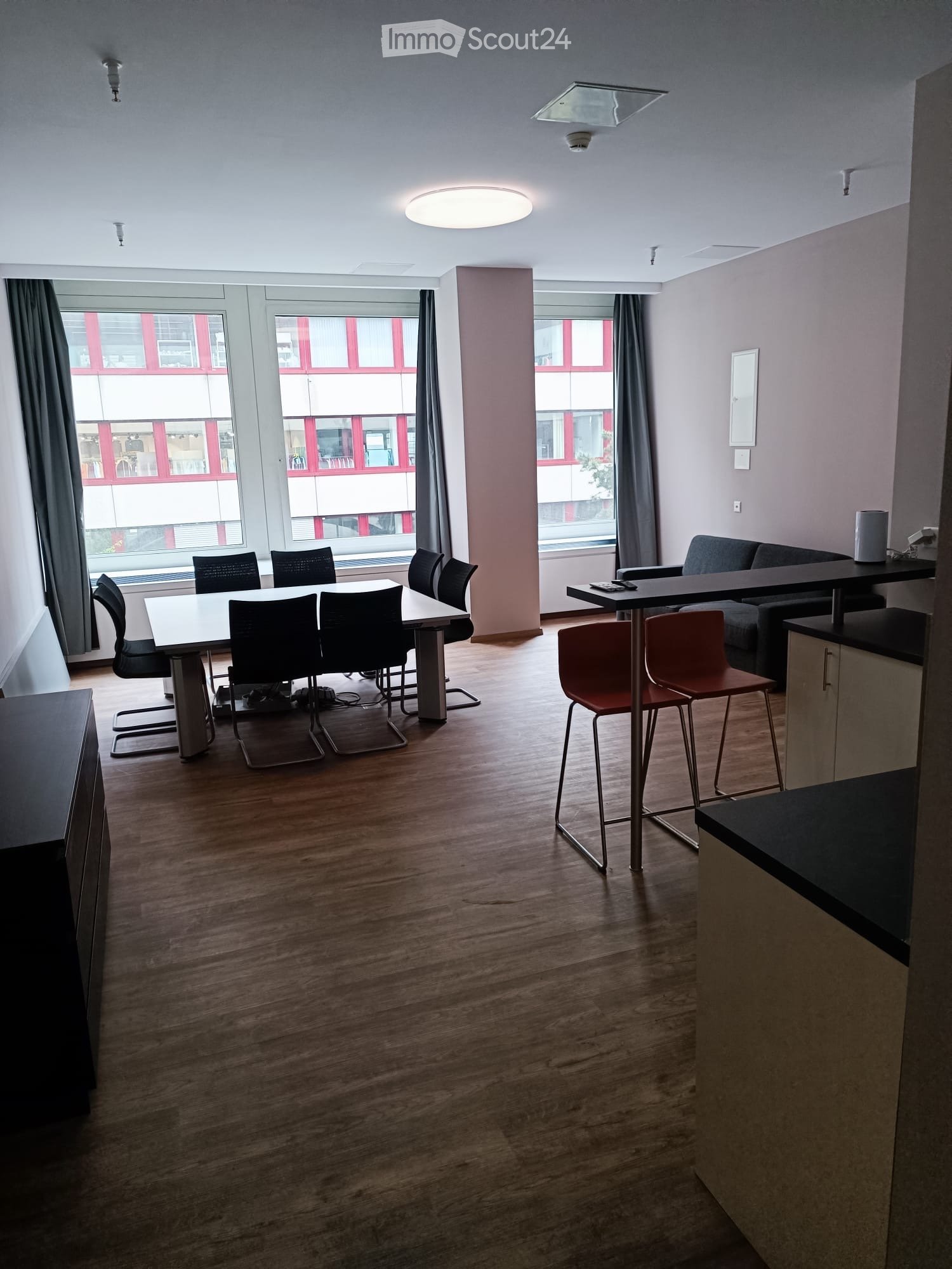 Open-plan room with wooden flooring, large windows, a sofa, a dining table with chairs, and a bar area with stools.