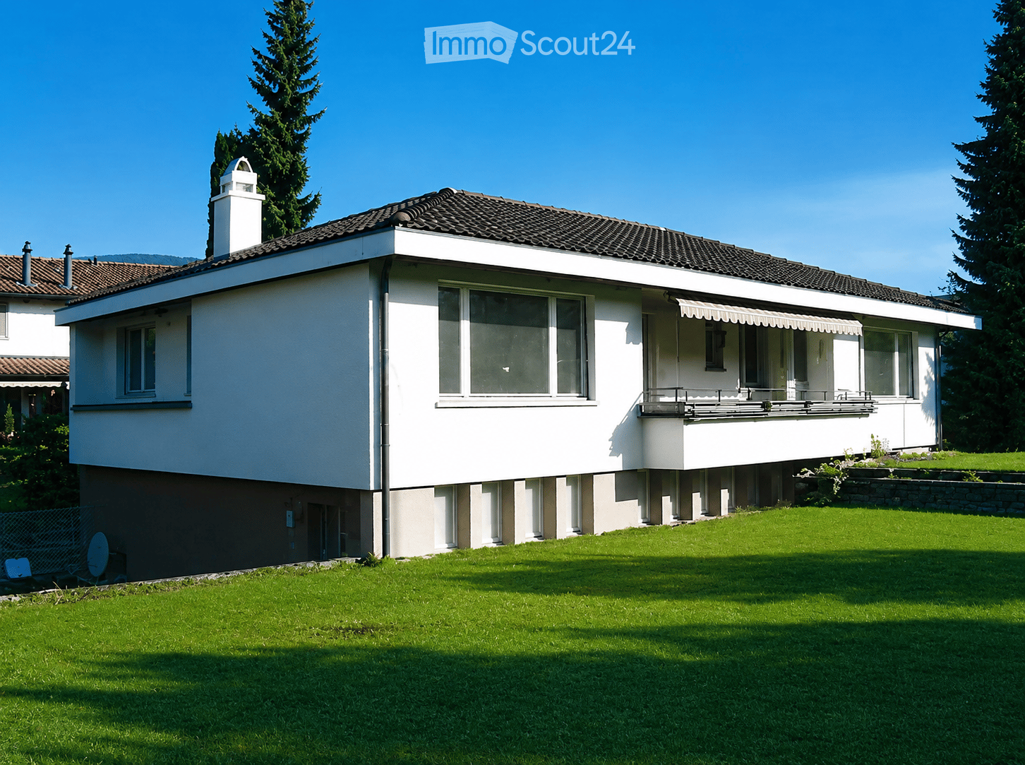 Two-story house with white exterior, dark tiled roof, multiple windows, and a covered balcony area. The house is surrounded by a well-maintained lawn and trees.
