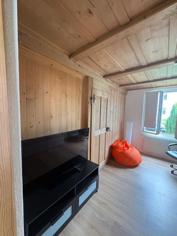 A modern living room with wooden walls and ceiling, featuring a large flat-screen TV on a black stand, an orange bean bag chair, and a black office chair near a window.
