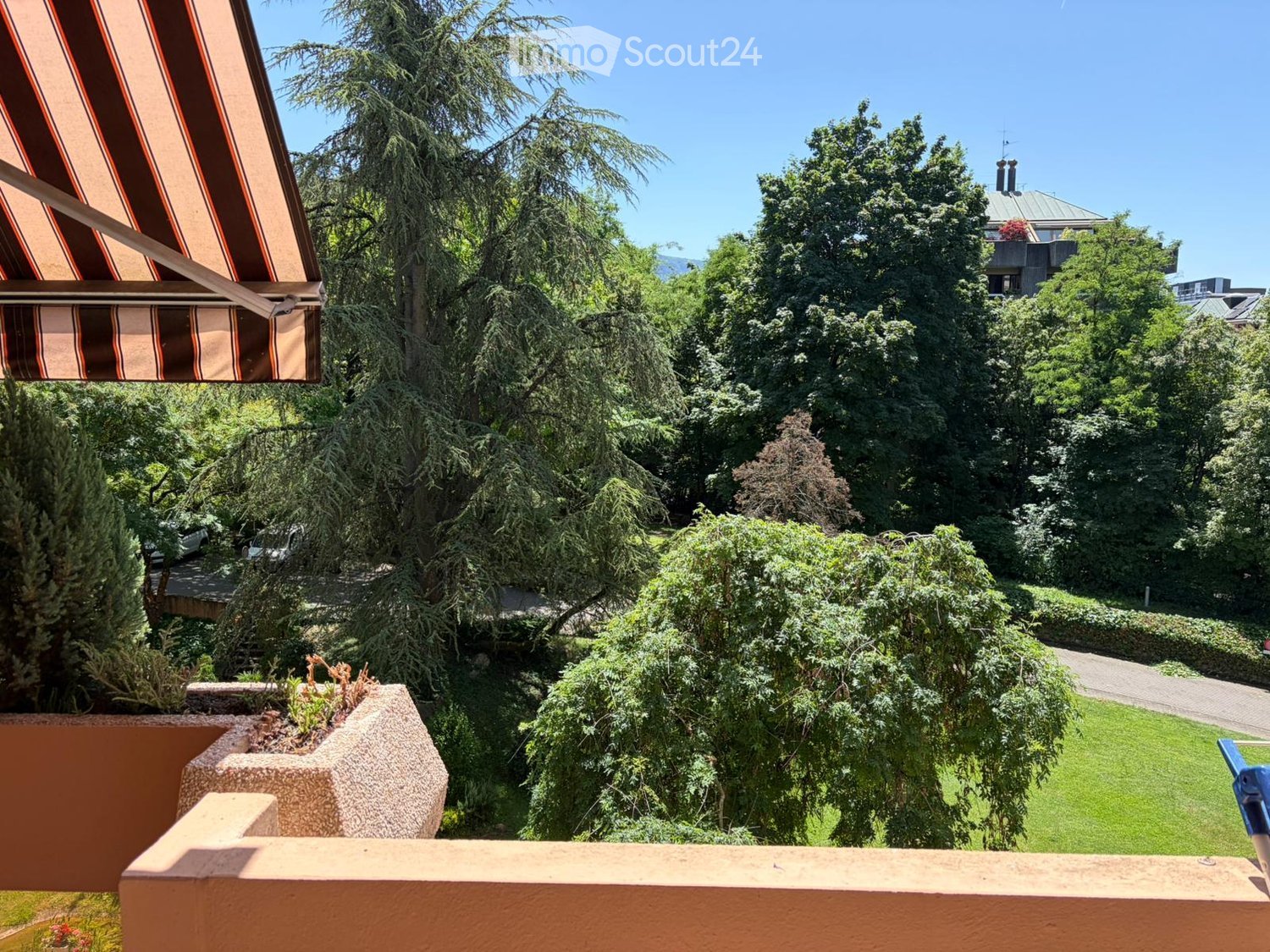 Balcony with wooden railing, overlooking a lush green garden and trees, with a striped awning on the left side.