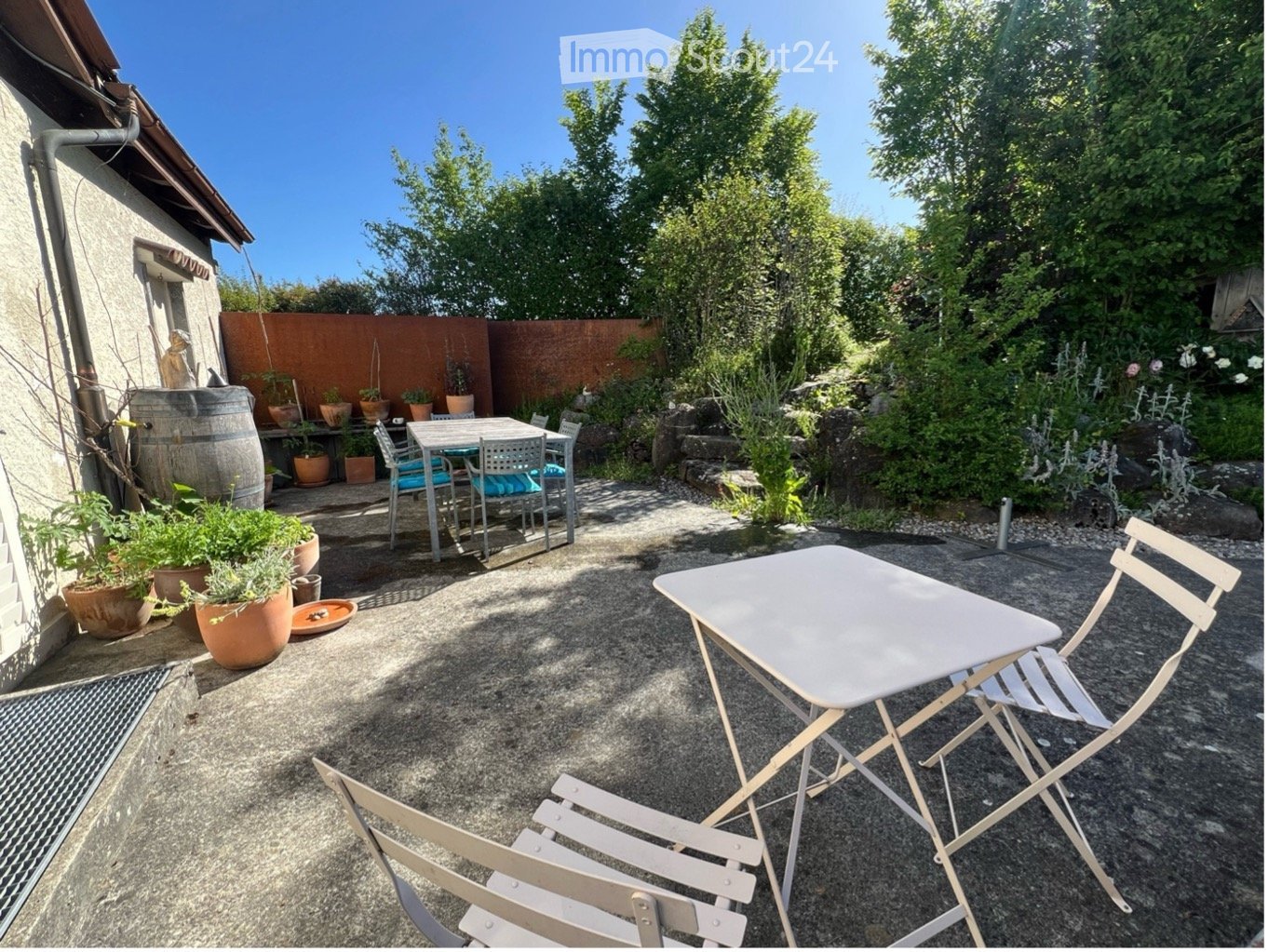 A well-maintained garden area with a gravel ground, featuring a wooden table and chairs, potted plants, and a wooden fence. The space is surrounded by lush greenery and trees, with a barrel and additional seating in the background.