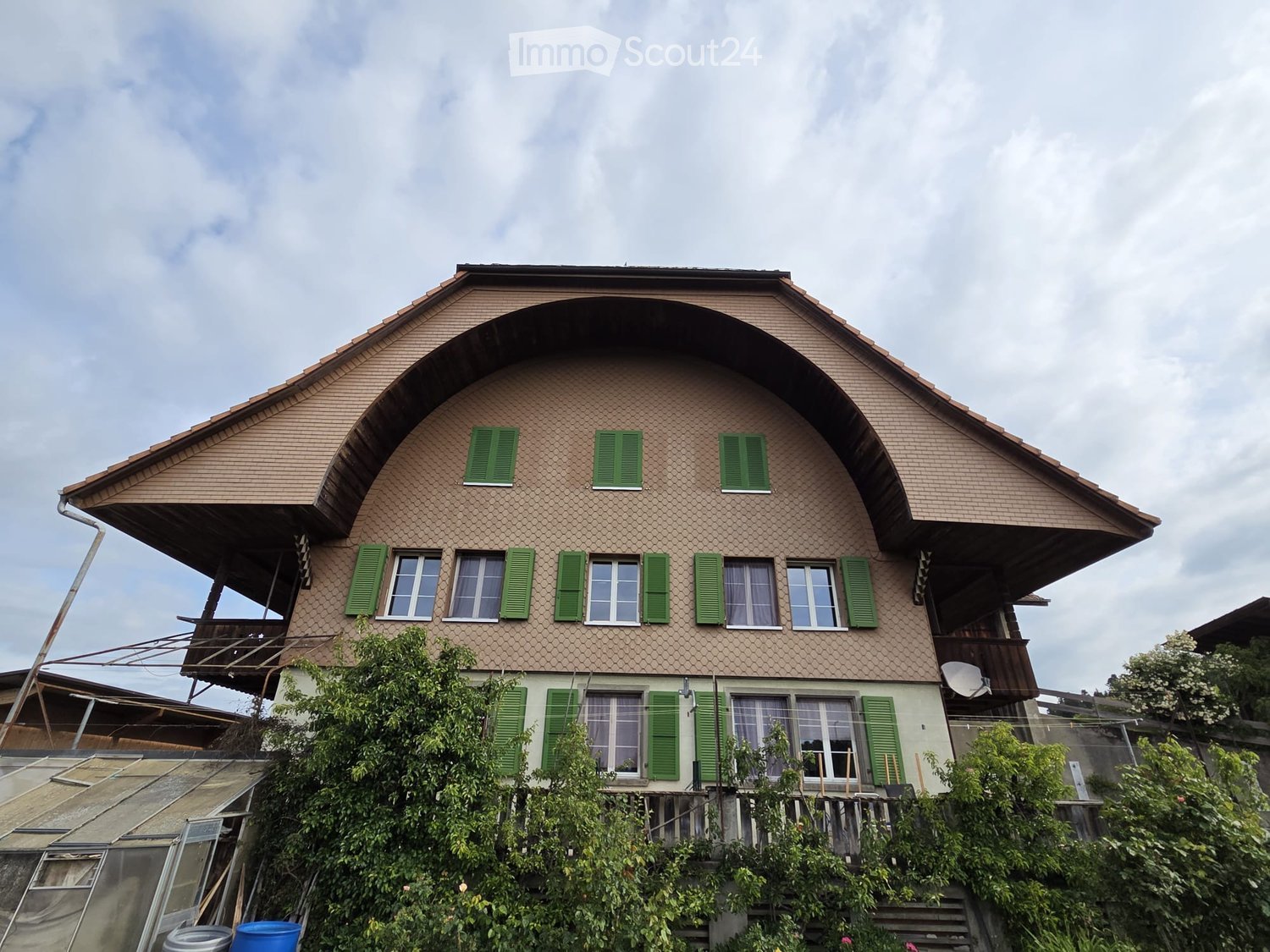 Two-story house with brown tiled roof and green shutters, surrounded by trees and a small garden area.
