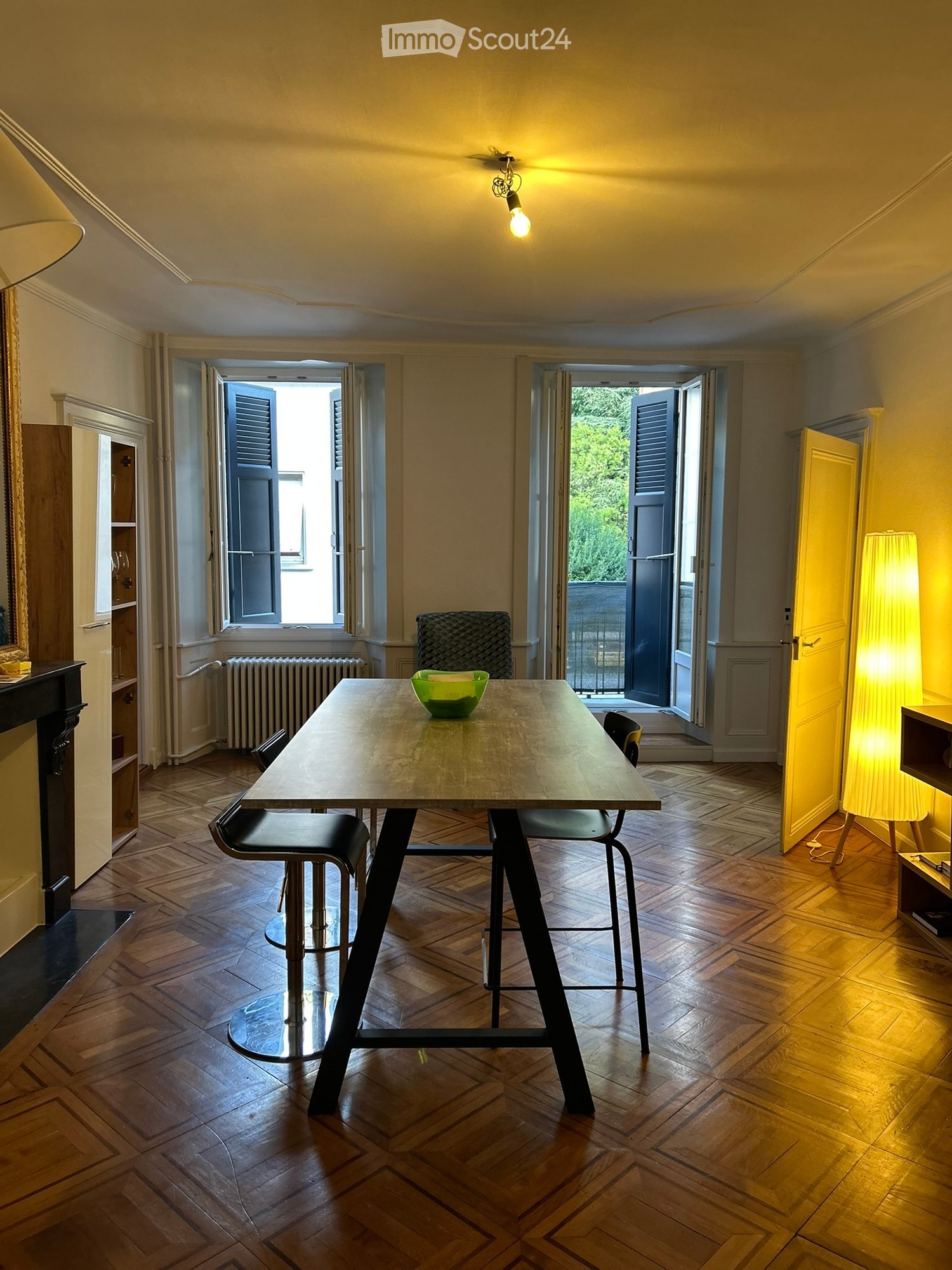 Dining area with wooden herringbone floor, modern table with four stools, green bowl on table, large windows with shutters, warm lighting from pendant lamp and floor lamp.
