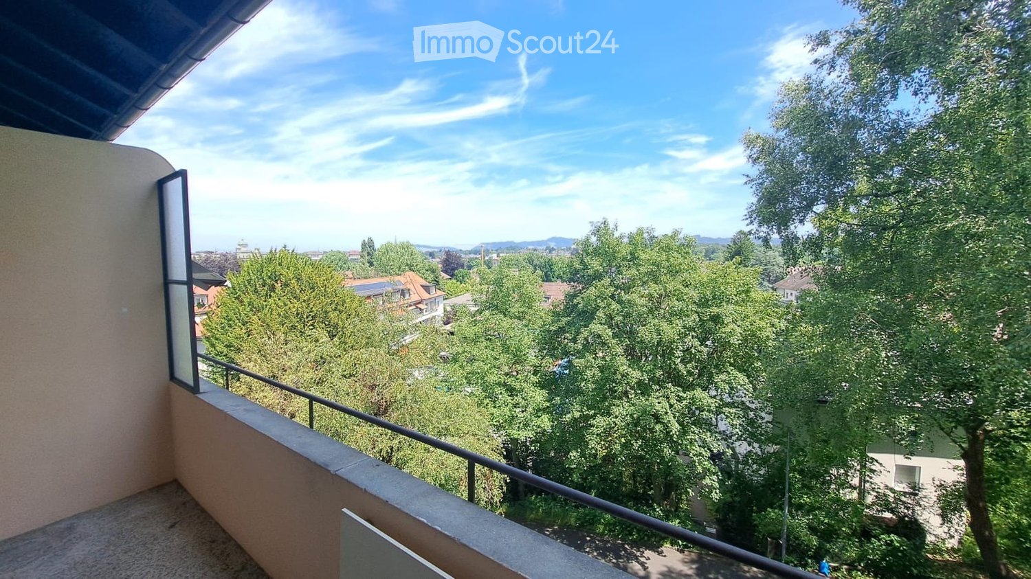 Balcony with metal railing, overlooking a tree-filled area and distant cityscape under a clear blue sky.