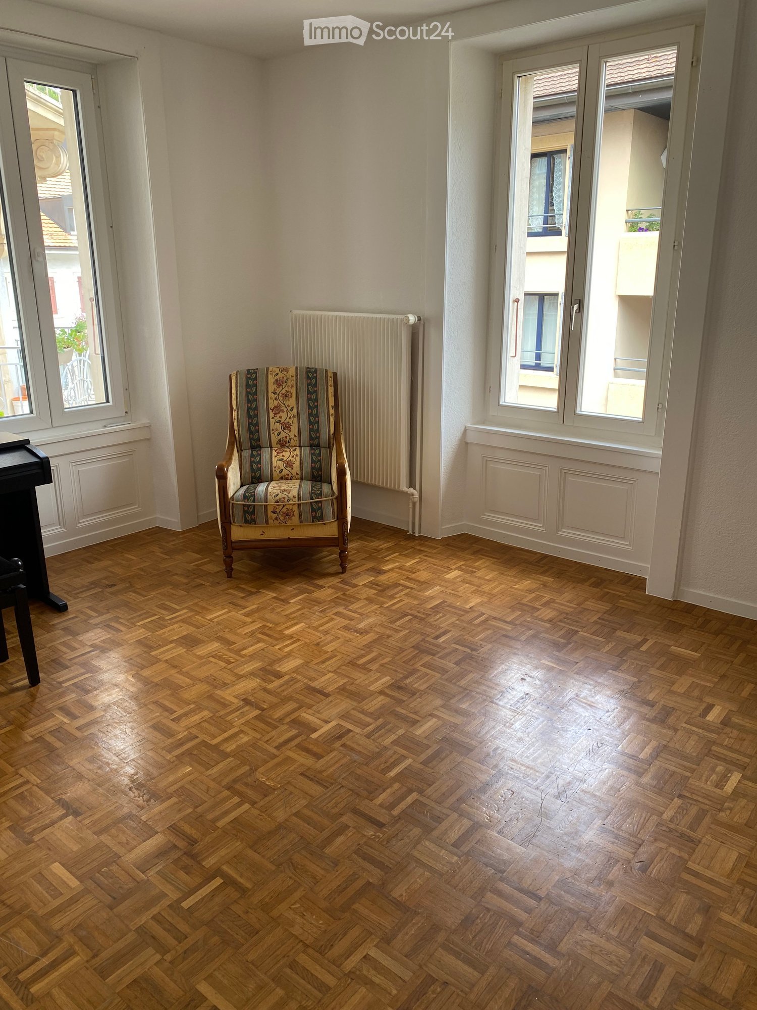 A small, well-lit room with wooden herringbone flooring, a single armchair with floral upholstery, and a radiator against the wall. Large windows offer views of the street and neighboring buildings.