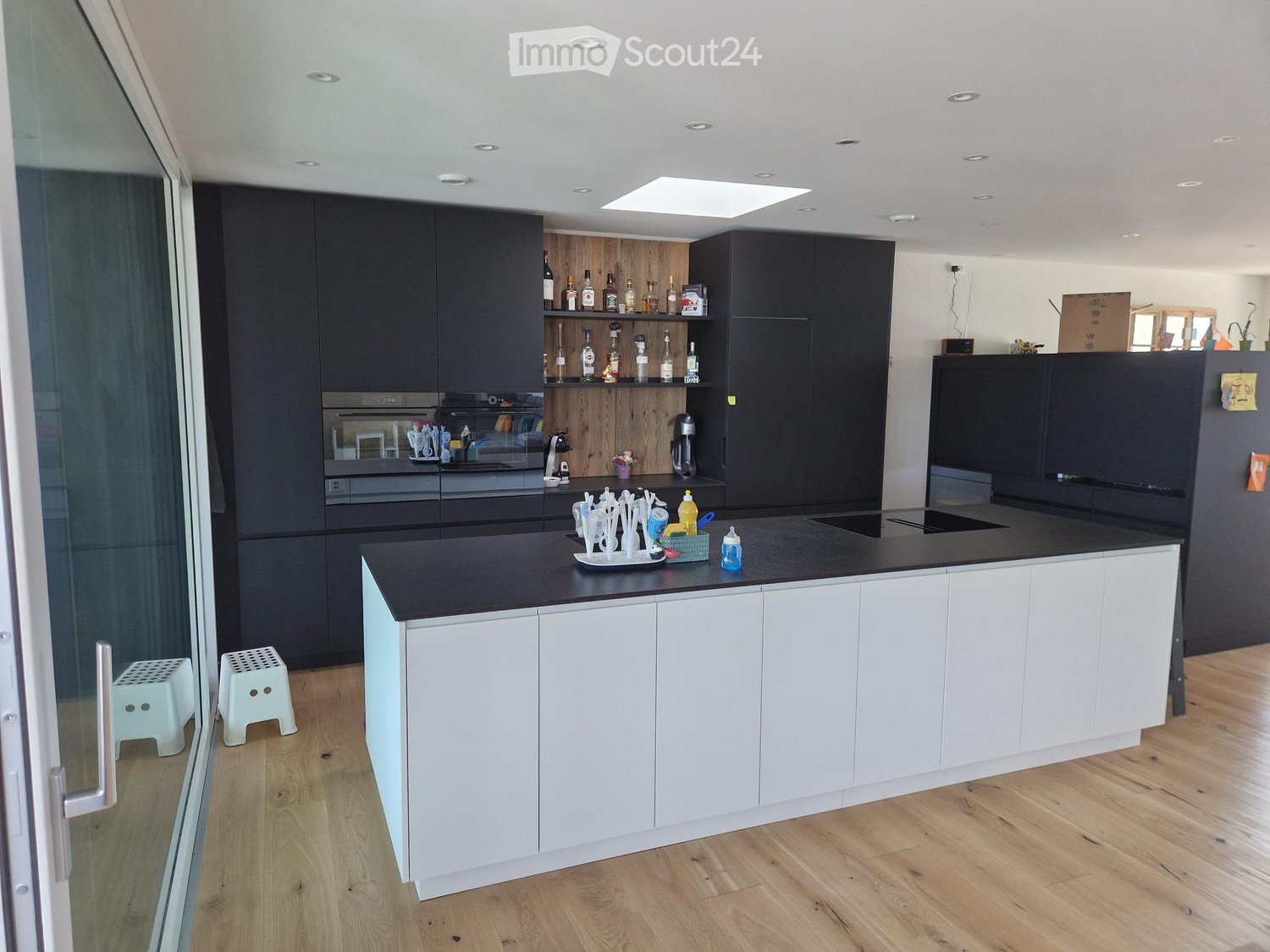 Modern kitchen with dark and white cabinets, black countertop, built-in oven, fridge, coffee maker, bottle rack, and two white stools near the window.