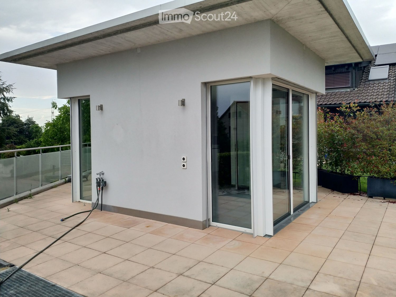 Modern balcony with large glass sliding doors, tiled floor, and a sleek white wall. Features a glass railing and outdoor lighting fixtures.