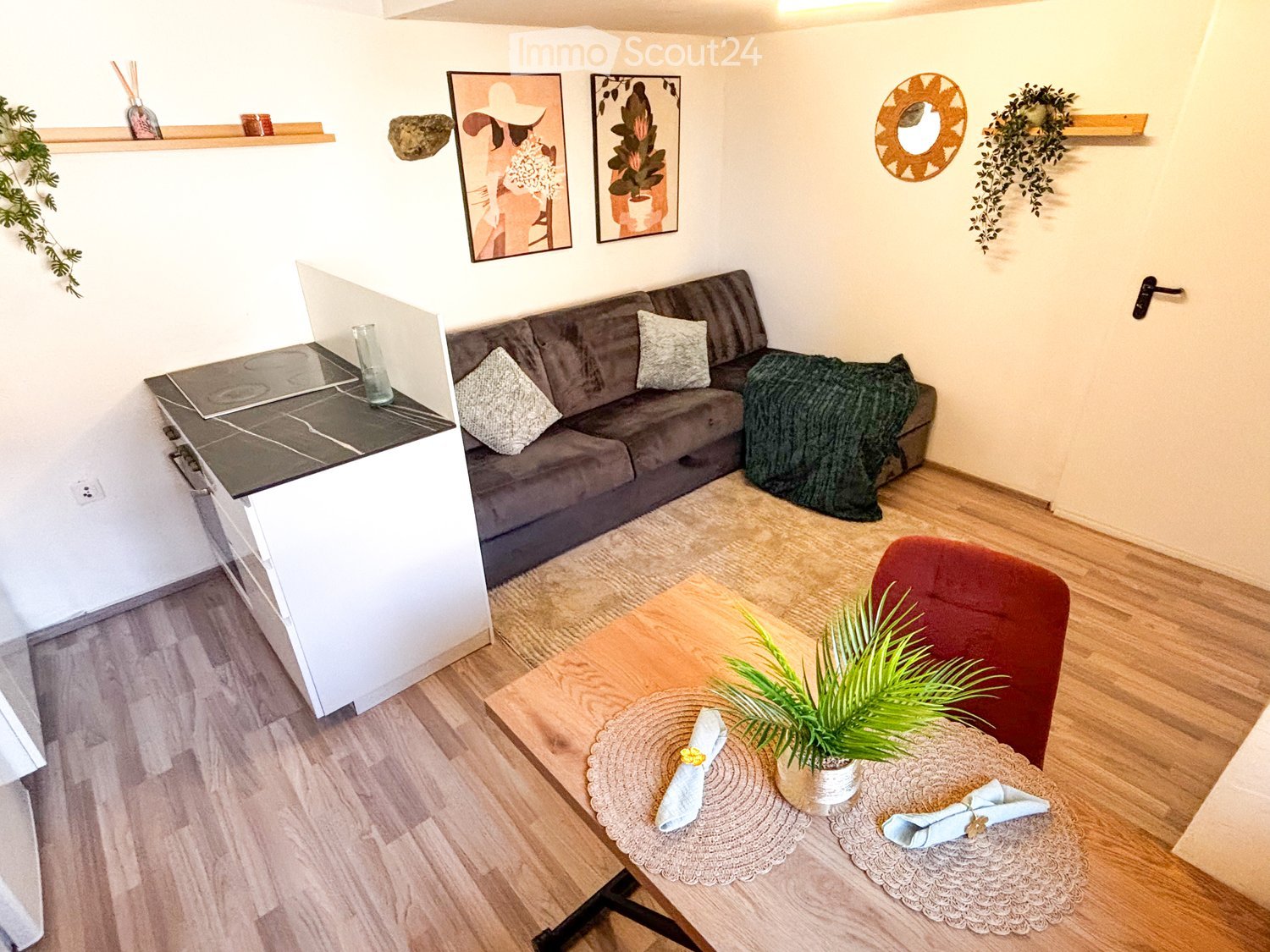 A modern, compact living space with a grey sofa, wooden coffee table, red chair, small kitchen counter, and decorative elements including a mirror and artwork.