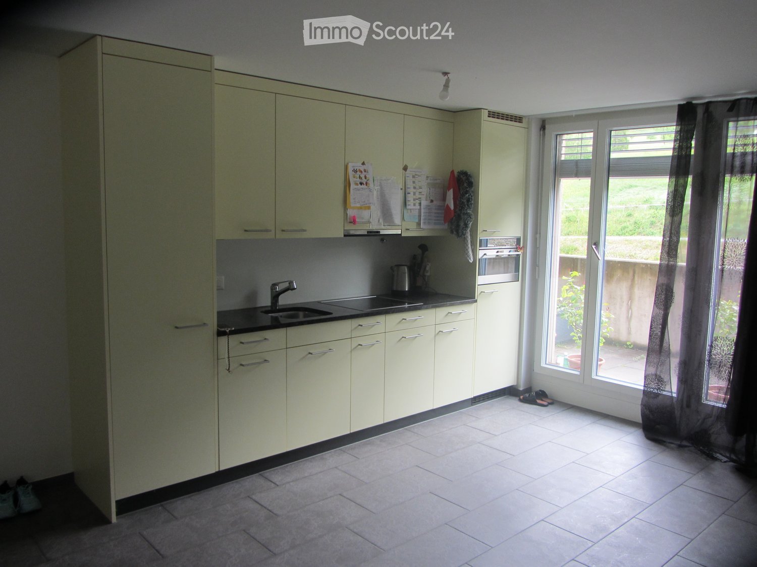 Kitchen. Modern kitchen with light-colored cabinets, a sink, an oven, and a window with a curtain. The floor is tiled, and there are some shoes near the window.