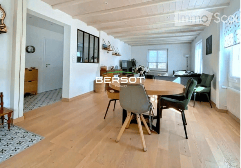 Bright, open-plan living and dining area with light wooden flooring, a round dining table with chairs, a sofa, a television, wall-mounted shelves, and a flower vase on a side table.