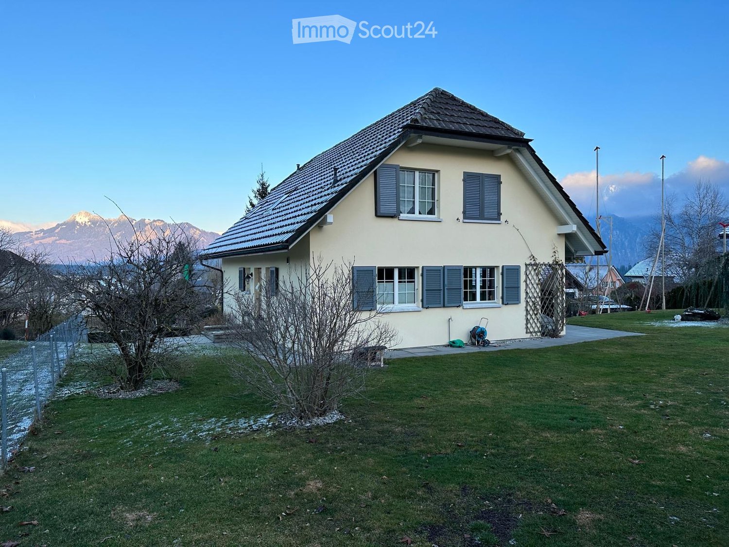 Two-story house with beige walls, dark blue shutters, and a tiled roof, surrounded by a grassy lawn and mountains in the background.