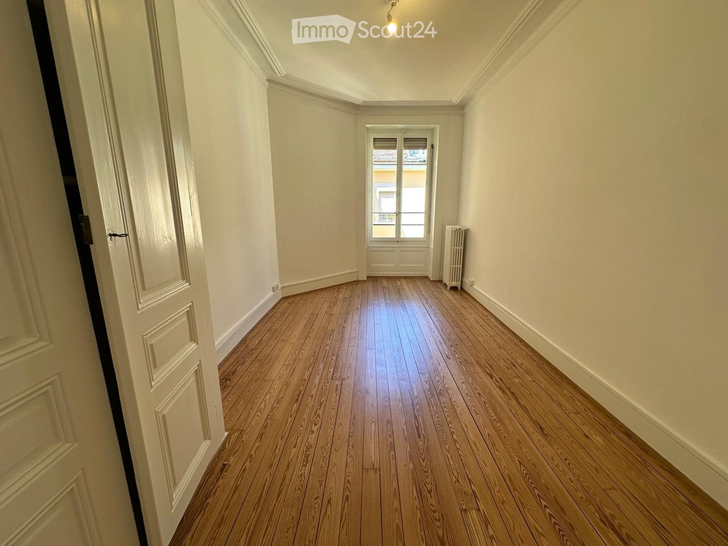 Empty hallway with wooden flooring, white walls, and a window at the end. A radiator is visible on the right side.