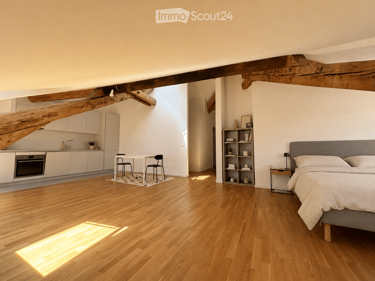 Open-plan living space with a bedroom and kitchen area. Features a wooden floor, exposed wooden beams, a grey upholstered bed, a nightstand, a small table with chairs, a kitchen with white cabinets, an oven, and a shelf with decorative items.