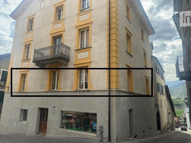 Multi-story building with yellow-framed windows and balconies, located in a European town with a visible street and storefront below.