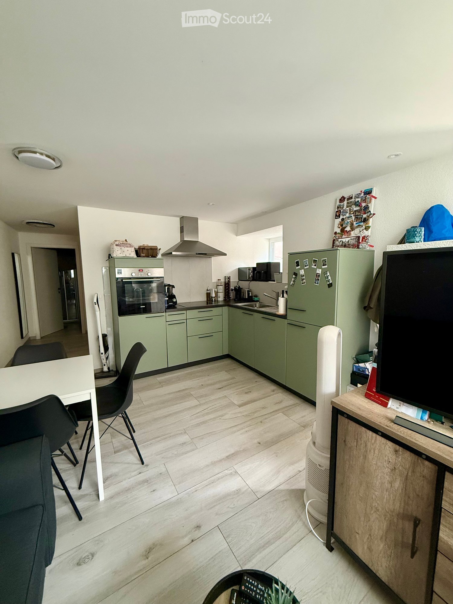 Open-plan living and kitchen area with light green cabinets, a white dining table with black chairs, a sofa, a television on a wooden stand, and a refrigerator with photos attached.