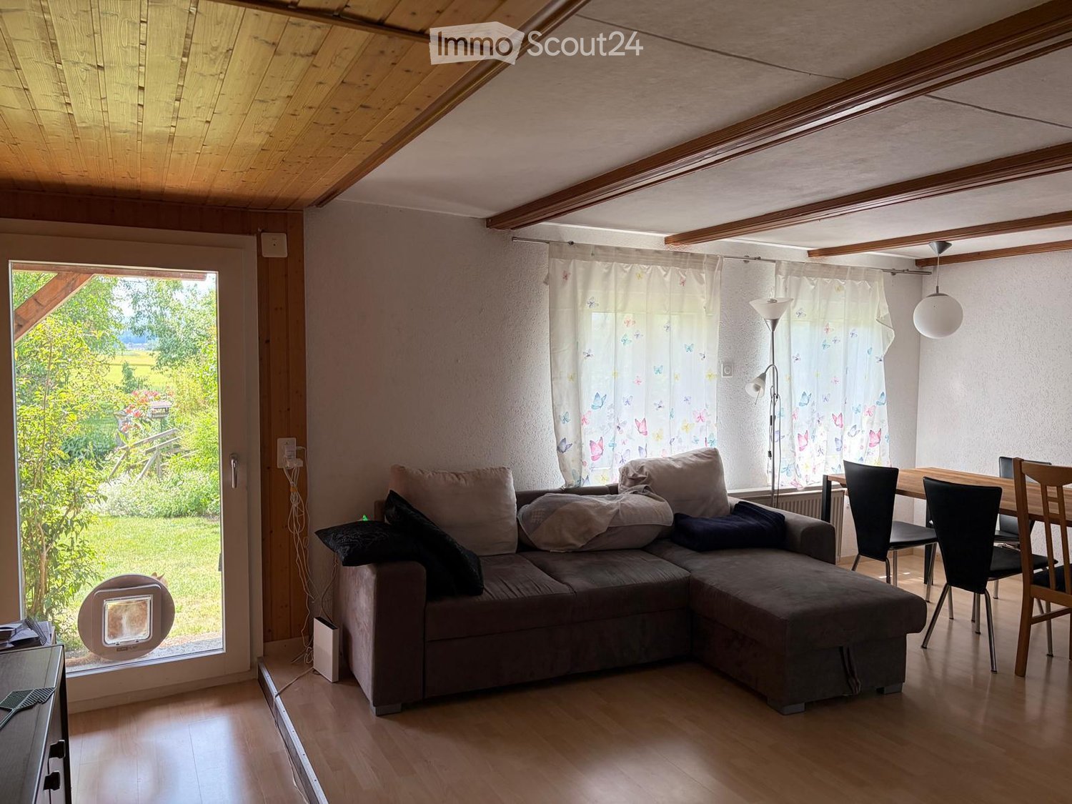 Bright living room with wooden floors, exposed wooden beams, large windows with sheer curtains, a grey sectional sofa, a dining table with chairs, and a floor lamp.