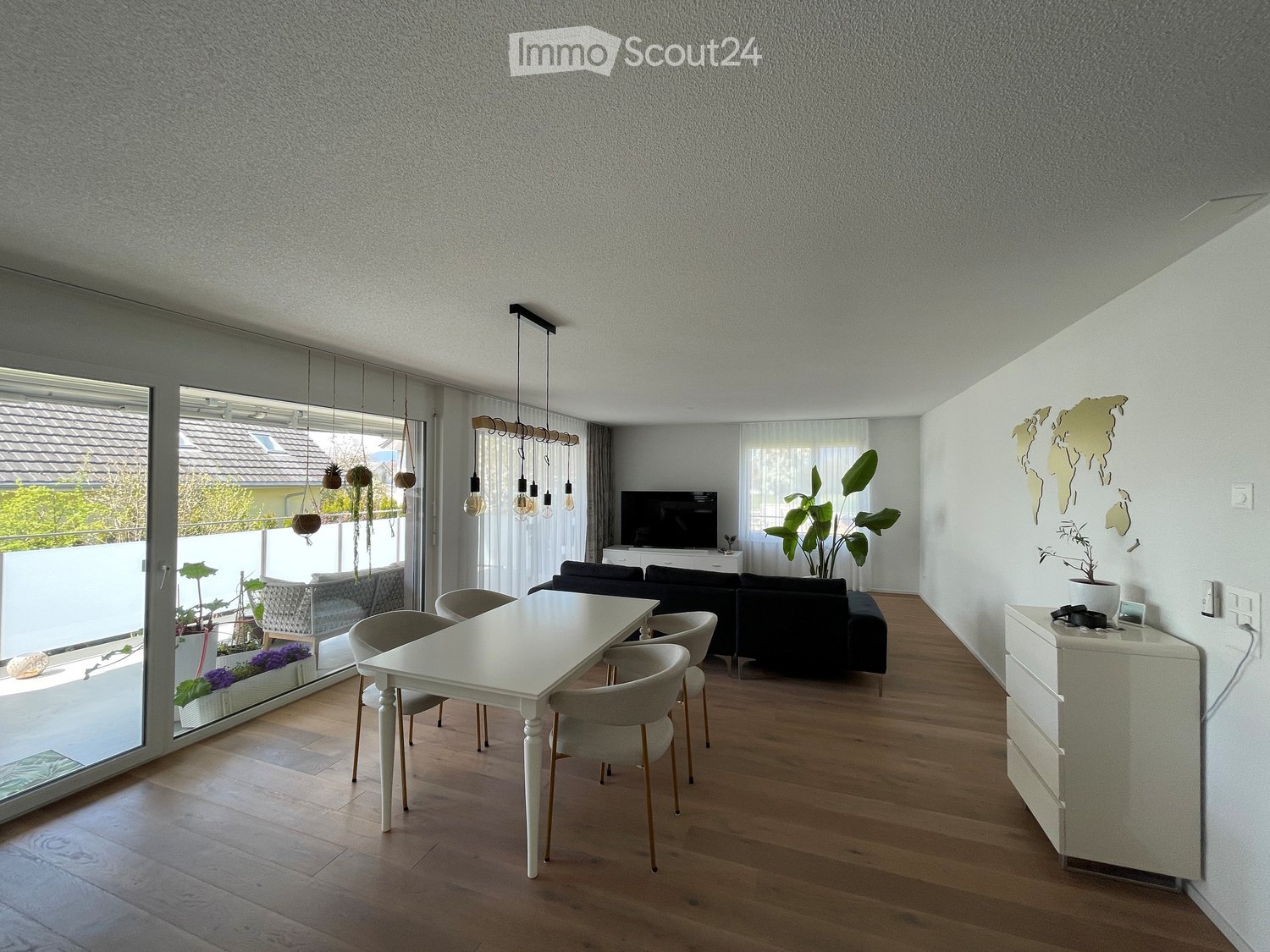 Modern living room with wooden flooring, large windows, a white dining table with chairs, a black sectional sofa, a TV, and decorative plants.