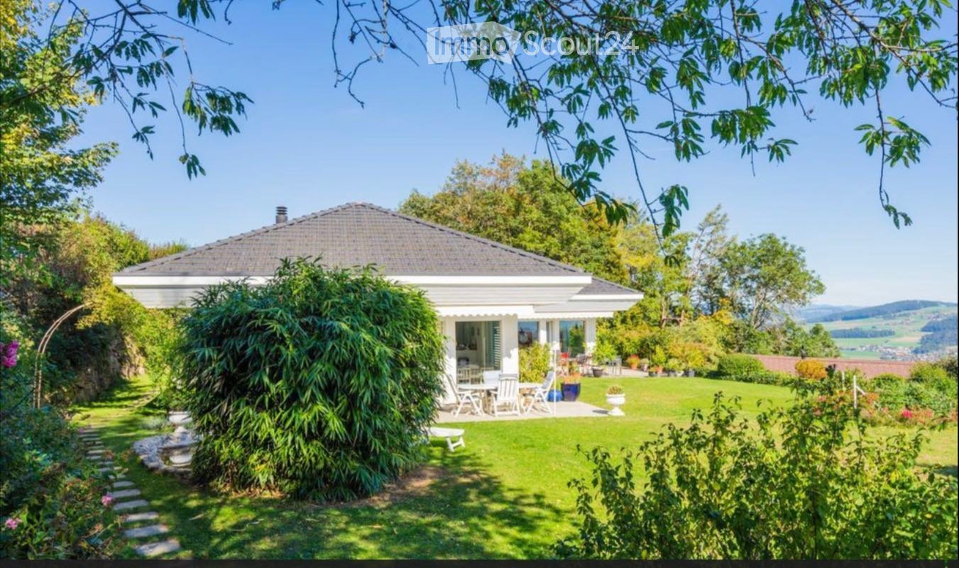 A white house with a tiled roof is surrounded by a well-maintained garden, featuring a patio area with outdoor furniture and a scenic view of the countryside.